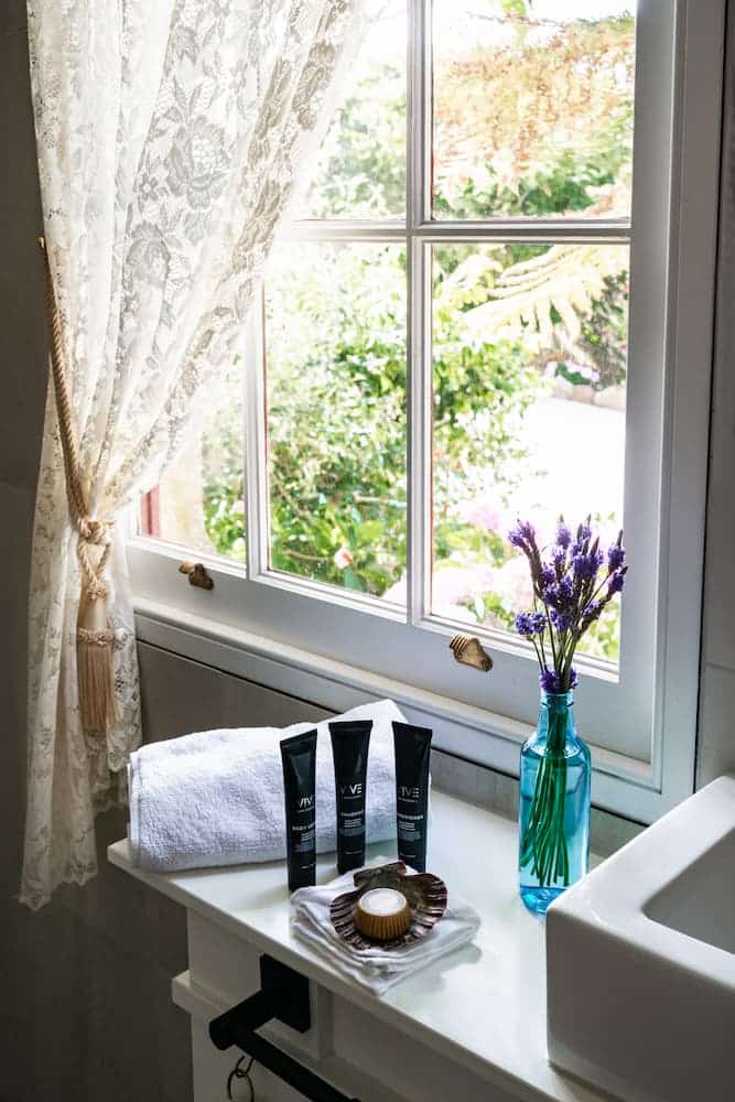 Bathroom countertop with folded white towel, three black toiletry tubes, shell-shaped soap dish, and blue vase with purple flowers by a window.
