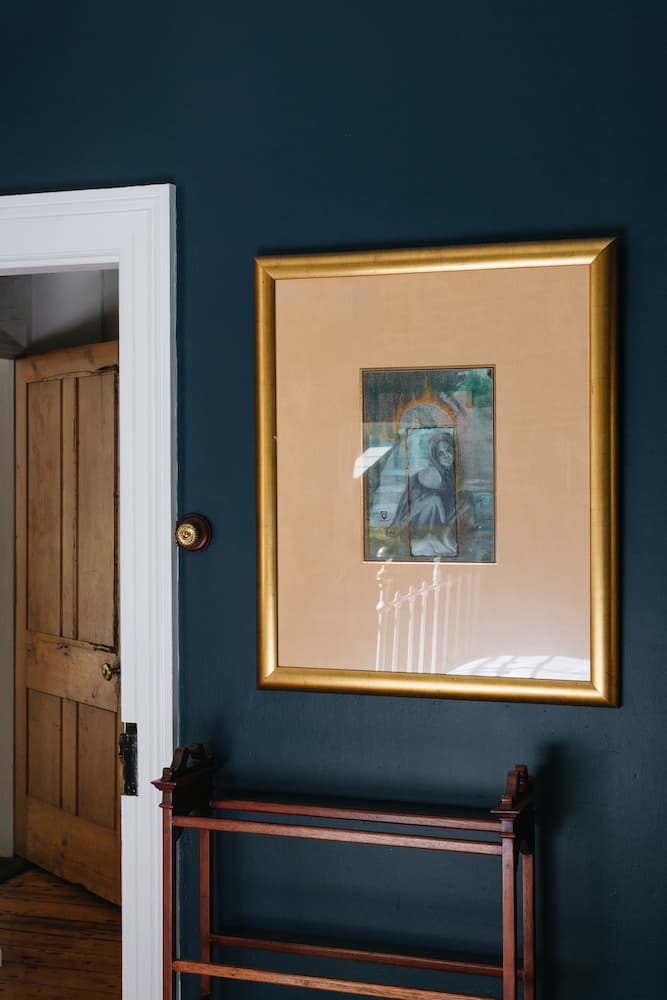 A framed painting with a gold border on a dark blue wall beside an open wooden door and a wooden rack below.