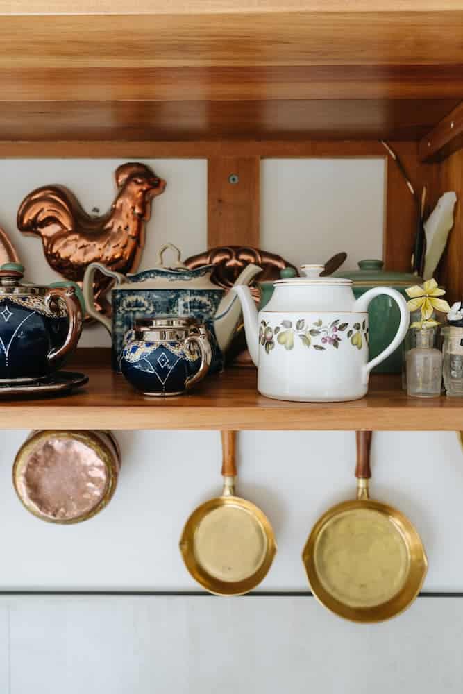 Wooden shelf with teapots and spice jars on top, copper moulds and brass pans hanging underneath.