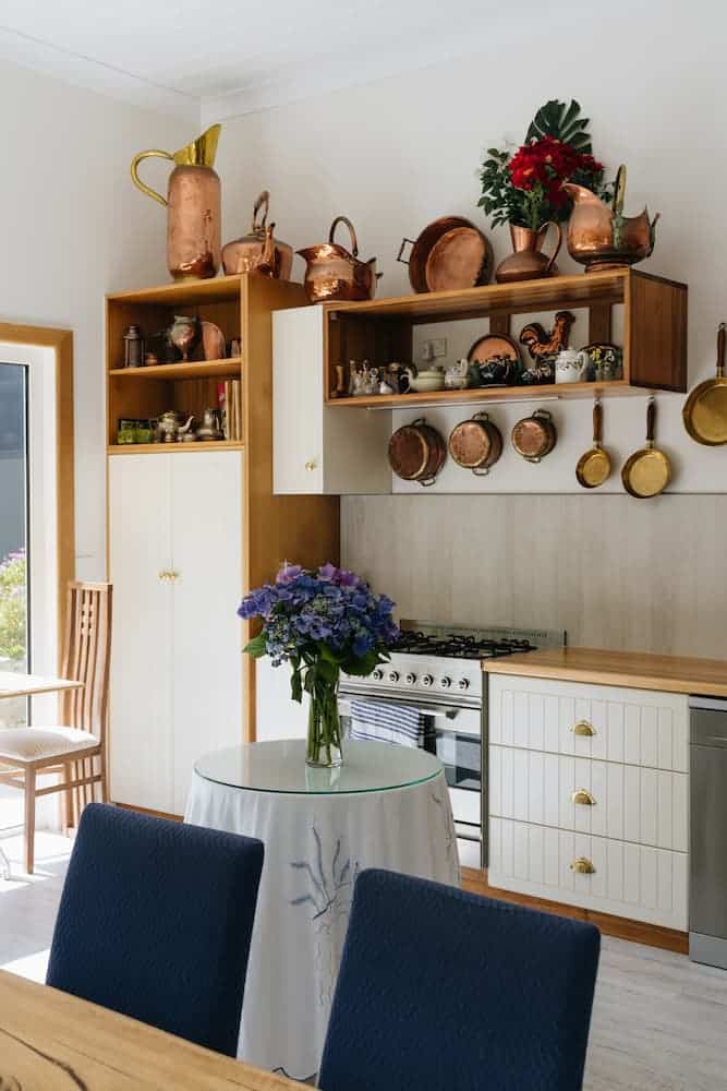 Kitchen with white cupboards, gold fittings, round table with flowers, blue chairs, shelves with copper cookware and red flowers.