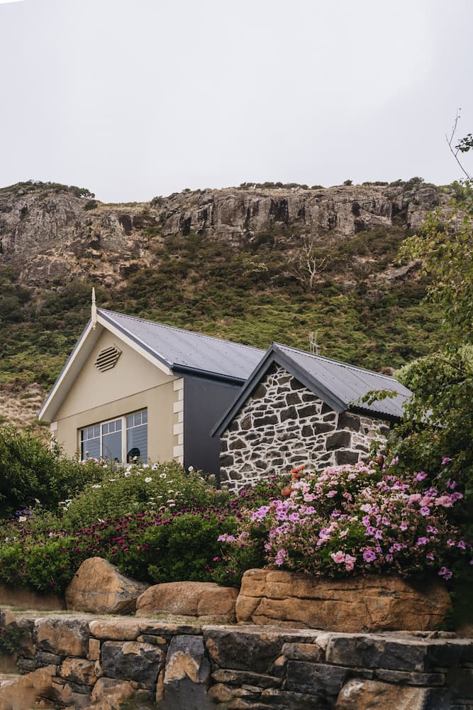 Stone and brick house with slate roof, flowering shrubs in front, rocky hillside in background.