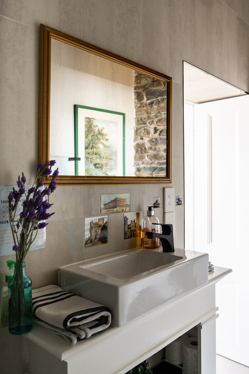 White rectangular basin with black tap under a large mirror; striped towels, toiletries, and purple flowers on the counter.