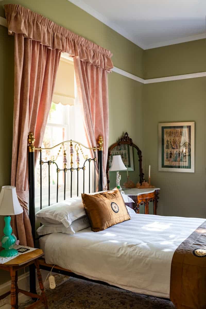 A bed with white linen and gold-brown accent cushions beside a window with pink curtains in a vintage bedroom with green walls.