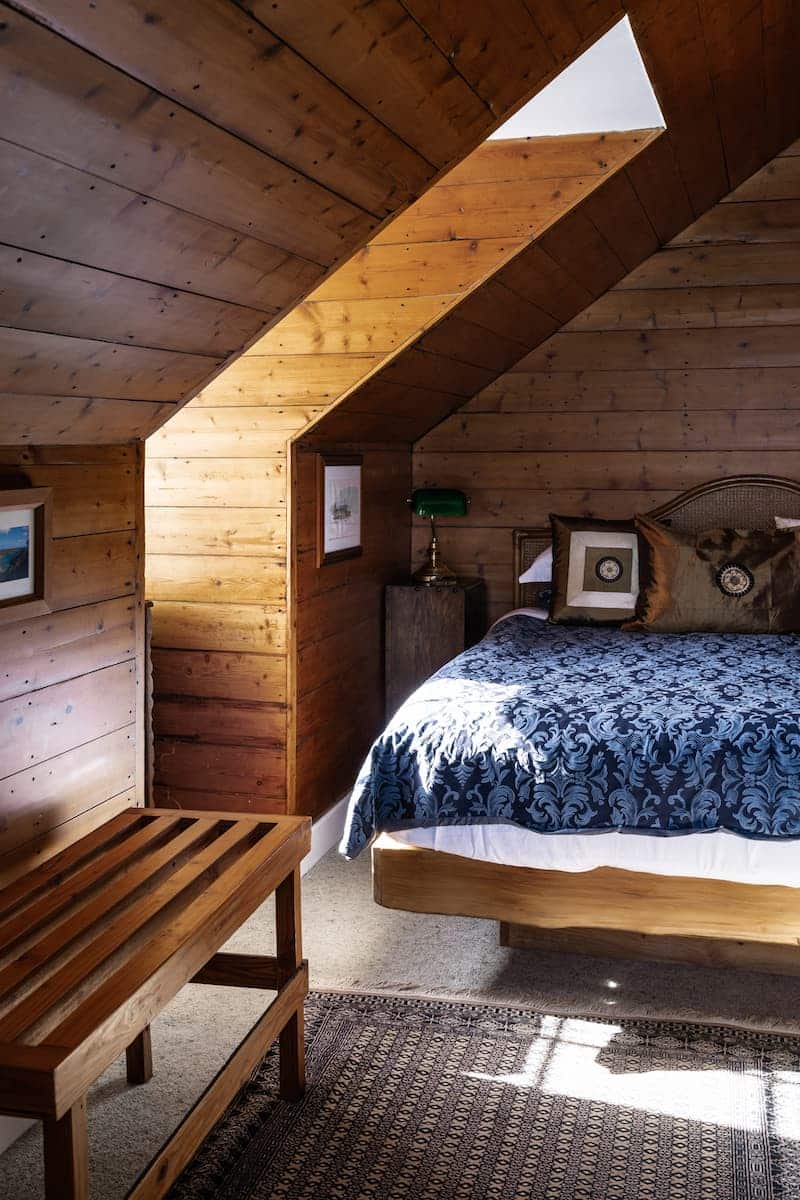Attic bedroom with wooden walls and ceiling, skylight, double bed with patterned bedlinen, framed art, wooden bench, bedside lamp.