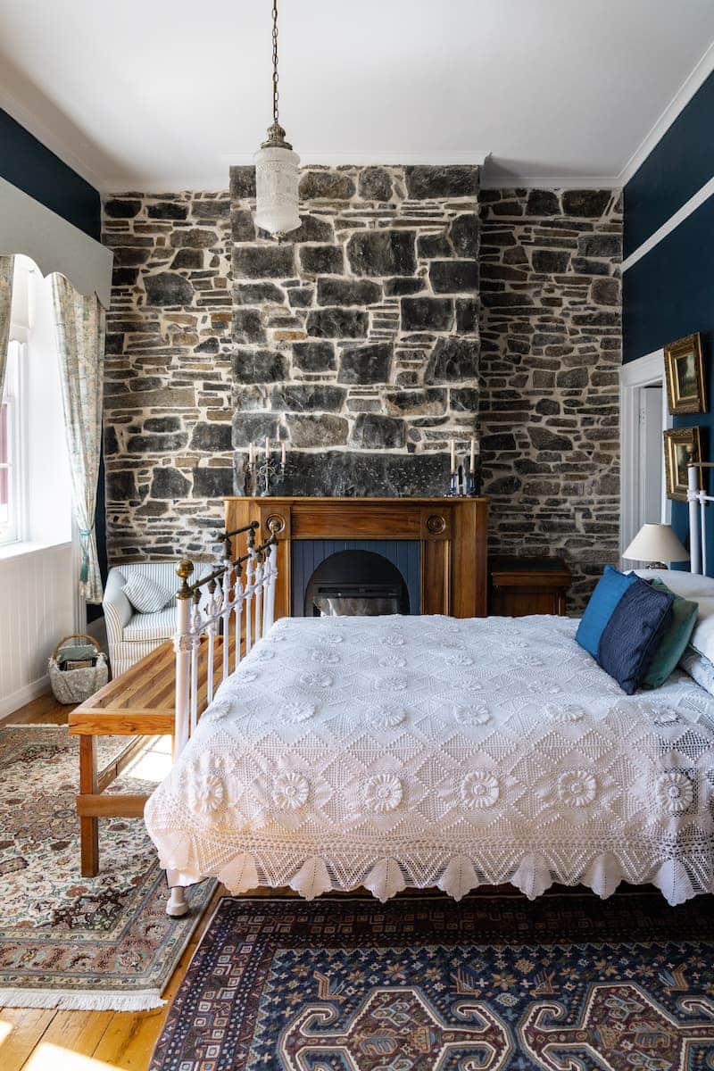 Stone fireplace, iron bed with white lace bedding, wooden bench, area rugs, light through curtained windows.