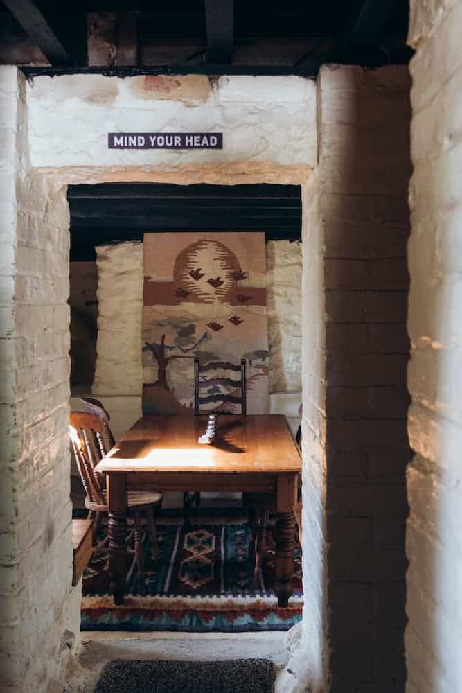 Wooden table with chairs in a small room with white brick walls, "Mind Your Head" sign above doorway, desert tapestry on wall.