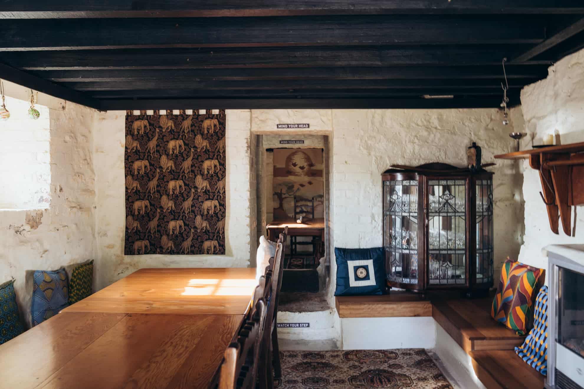 Long wooden table with wooden chairs, elephant tapestry on white brick wall, glass-fronted cupboard, and upholstered bench.