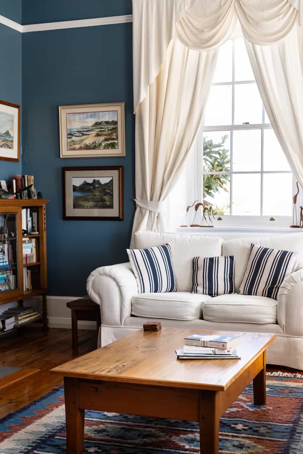White sofa with striped cushions, wooden coffee table, books, wall art, blue walls, bookcase, large window with cream curtains.